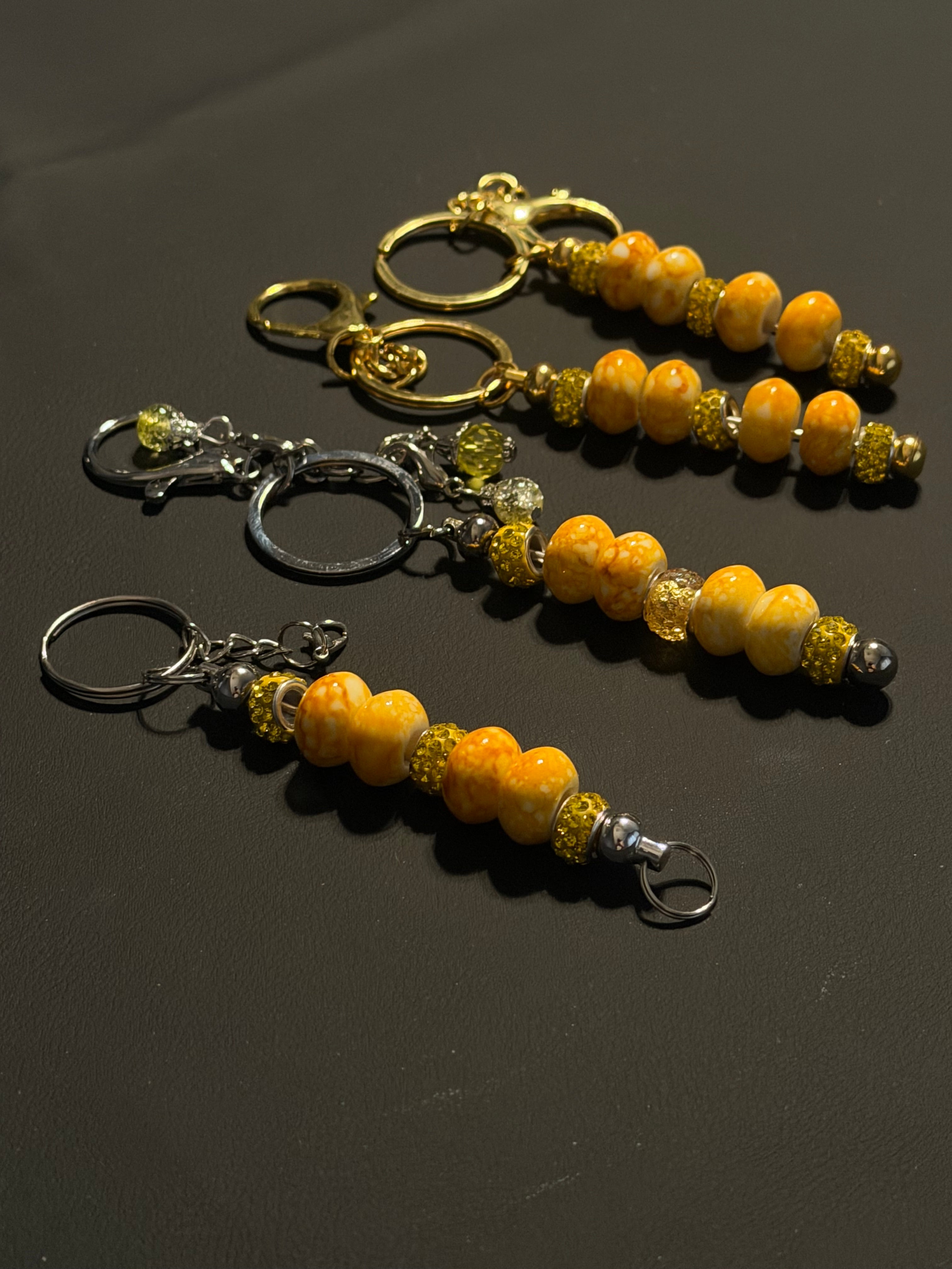 Three beaded keychains with orange and yellow marbled beads, metallic spacer beads, and split rings in gold and silver, displayed on a dark gray background.