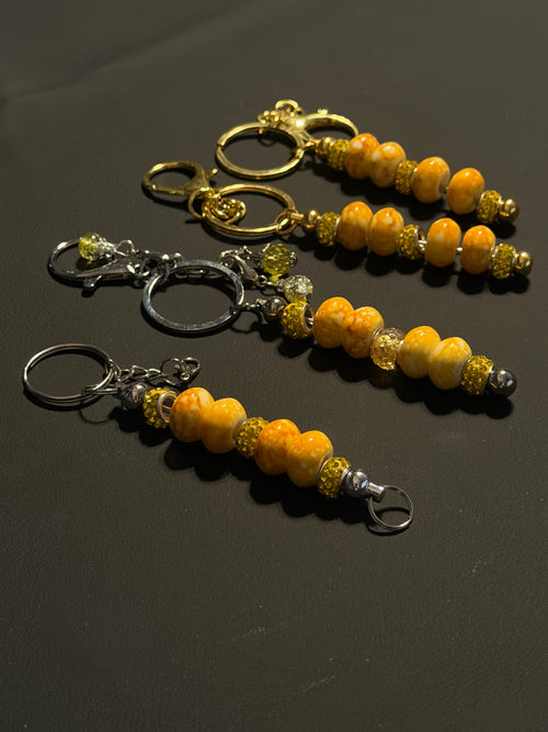 Three beaded keychains with orange and yellow marbled beads, metallic spacer beads, and split rings in gold and silver, displayed on a dark gray background.